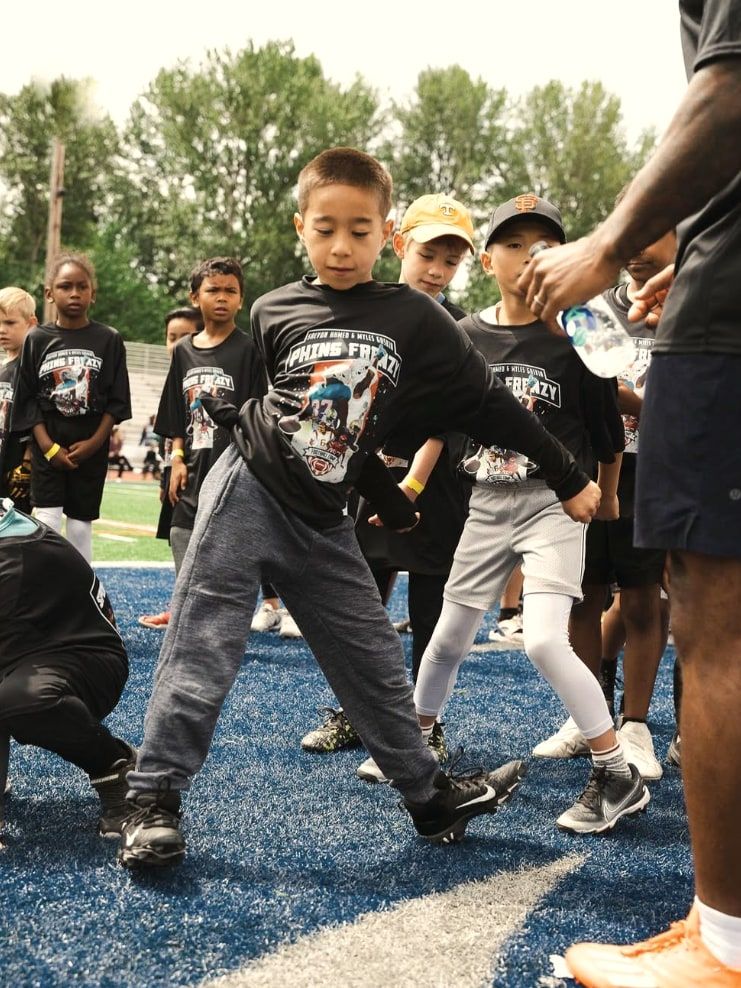 A boy in a camp shirt drops into a stance at the front of a drill line, a coach's hand holding a water bottle at the edge of the frame