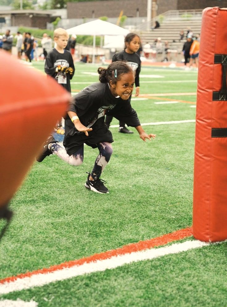 A girl in a camp shirt sprints past a red tackle pad, grinning, the stands behind her
