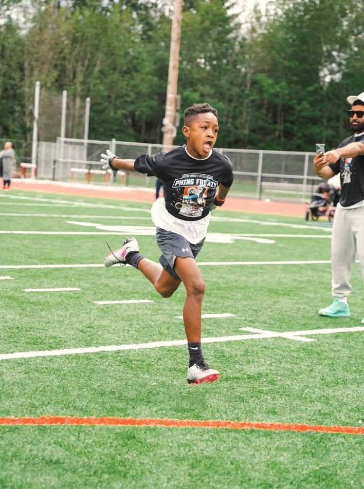 A boy runs a route down the field with his arms out, a parent filming on a phone behind him