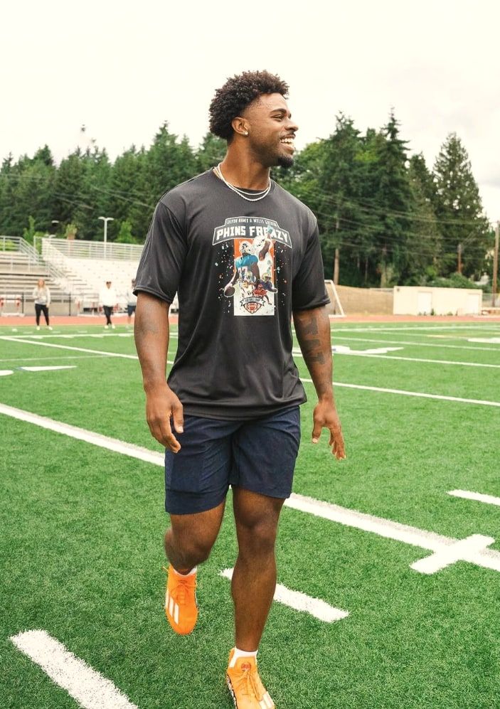 Salvon Ahmed walks the field in a camp shirt, laughing, the bleachers and the trees behind him