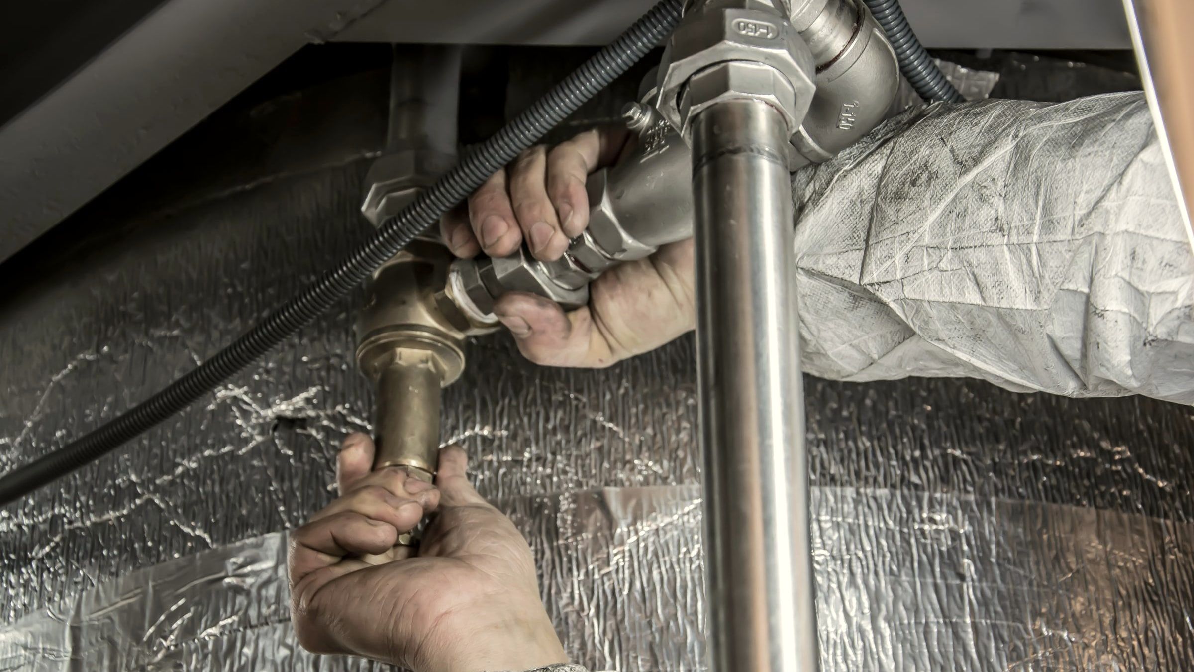 Two bare hands fitting a steel pipe joint in a cramped space under a sink, foil insulation behind the pipes